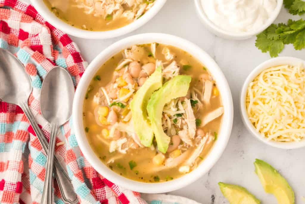 Two bowls of White Chicken Chili with avocado and cheese toppings, surrounded by small bowls of sour cream and shredded cheese.