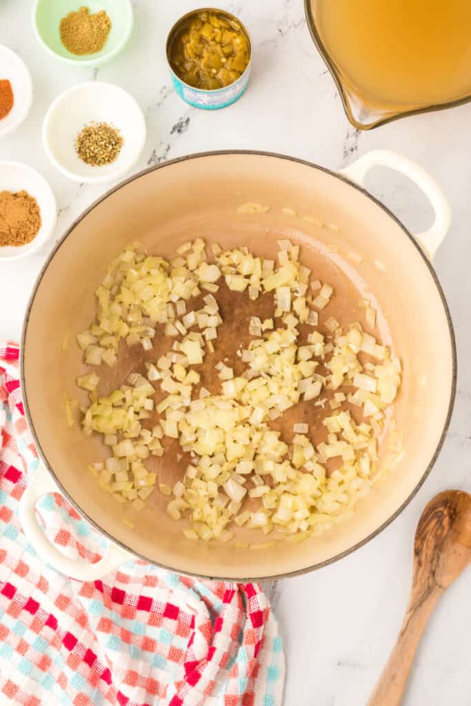 Onions and minced garlic sautéing in olive oil in a dutch oven with a colorful red and aqua towel beside it.