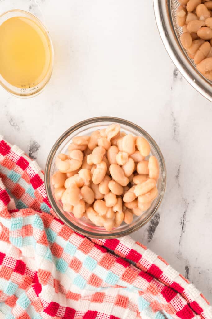 Bowl of rinsed white beans sitting beside a measuring cup of broth on a checkered towel.