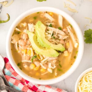 Square crop of a bowl of White Chicken Chili with avocado slices and cheese on top, served on a red and aqua towel with cilantro garnish.