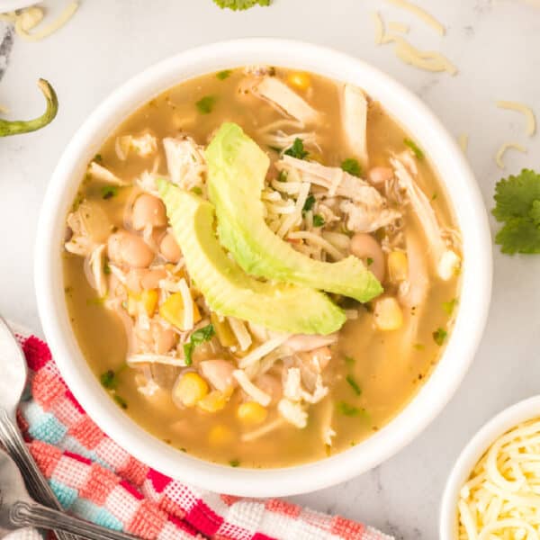 Square crop of a bowl of White Chicken Chili with avocado slices and cheese on top, served on a red and aqua towel with cilantro garnish.