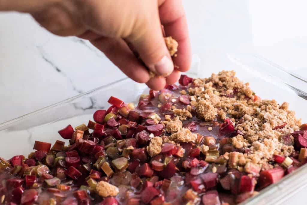 Hand sprinkling oat crumb mixture over rhubarb and sugar sauce in a baking dish.