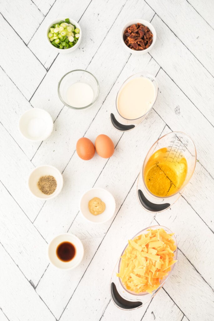 small bowls of chopped green onions, bacon pieces, spices, mustard, Worcestershire sauce, milk, beer, shredded cheese, and two eggs on a white wood surface