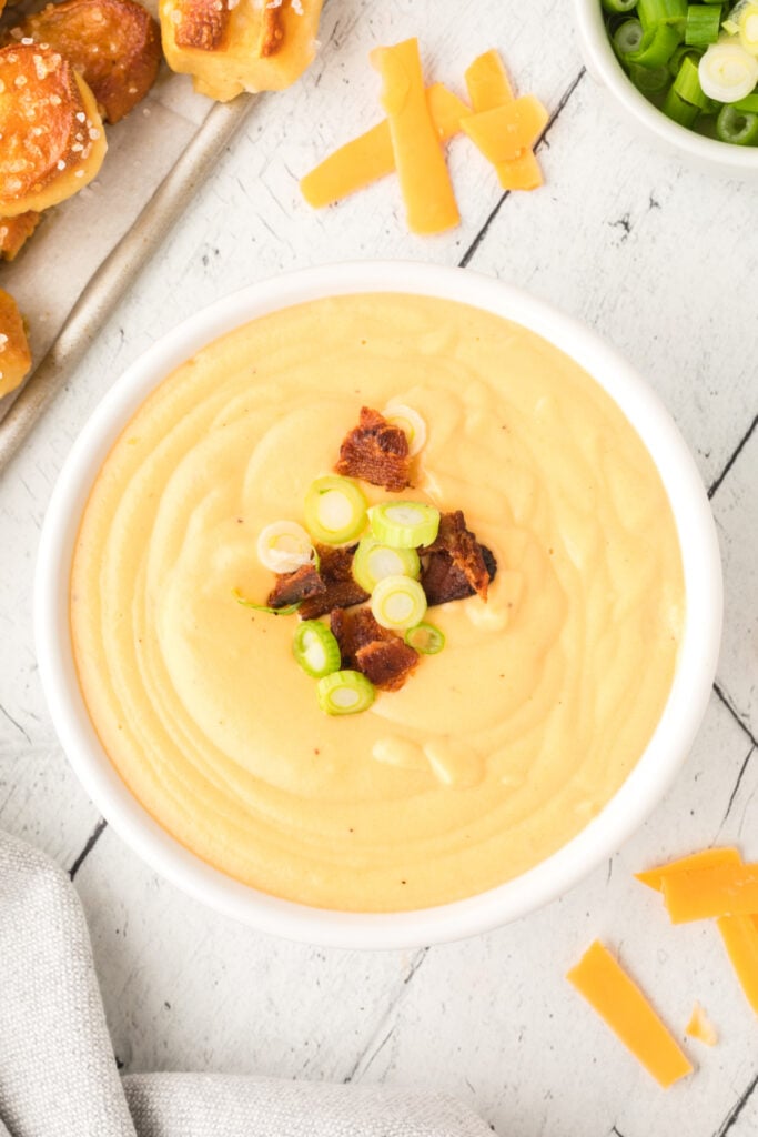 bowl of beer cheese dip topped with bacon pieces and sliced green onions with pretzels nearby
