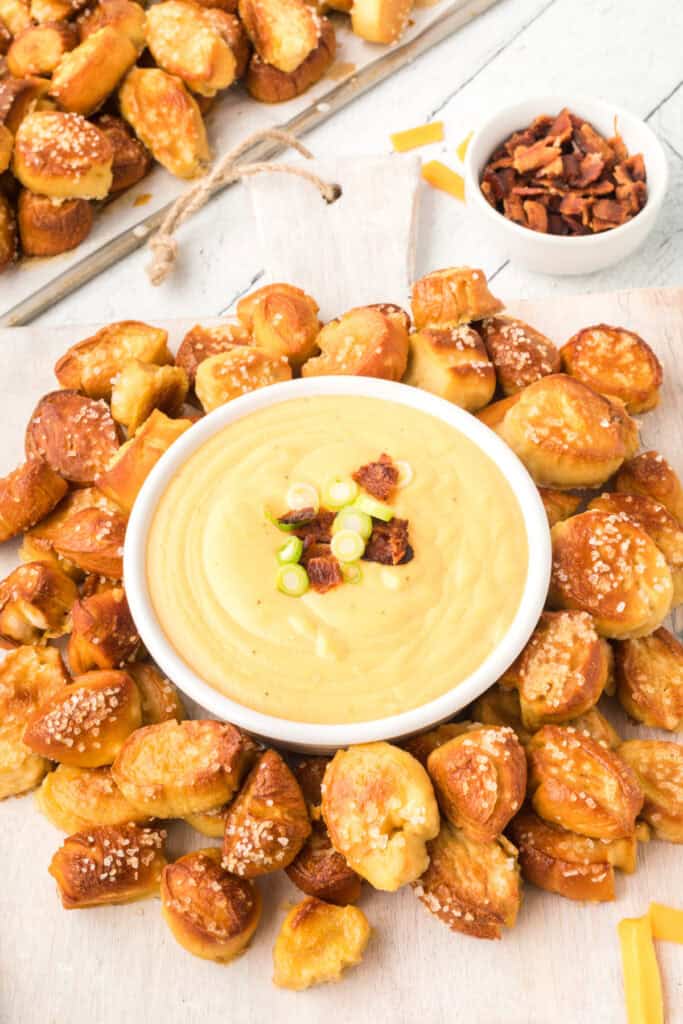 bowl of Beer Cheese Dip topped with bacon and sliced green onions surrounded by pretzel bites on a wooden board