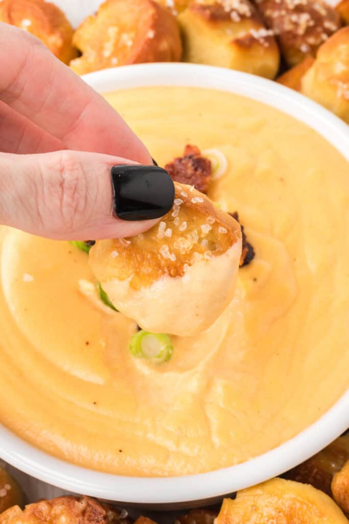 hand holding a pretzel bite dipped into Beer Cheese Dip with bacon and green onions on top