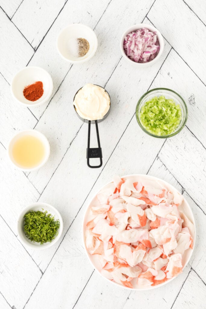Ingredients for Deli-Style Crab Salad including imitation crab, celery, red onion, dill, mayonnaise, lemon juice, and seasonings arranged on a white background.