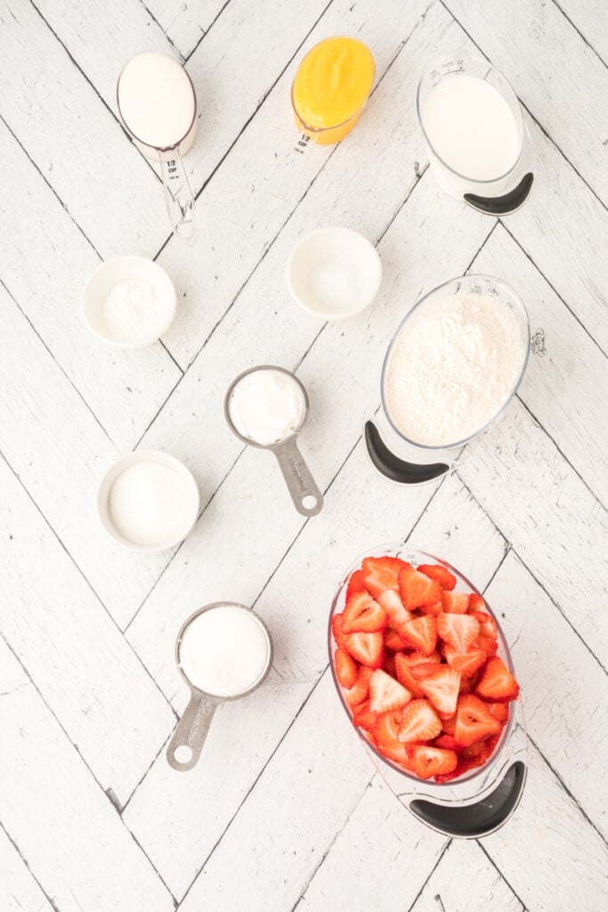 Measuring cups and bowls filled with ingredients, including sliced strawberries, sugar, flour, and lemon curd.