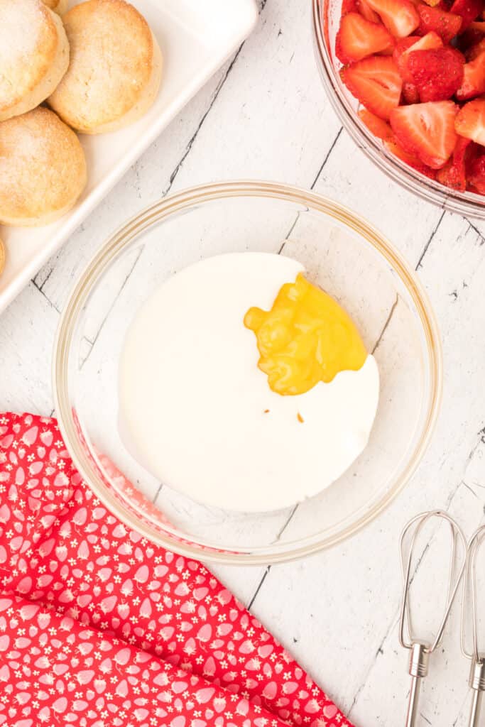 Bowl of cream with a dollop of lemon curd on top, strawberries and biscuits nearby.