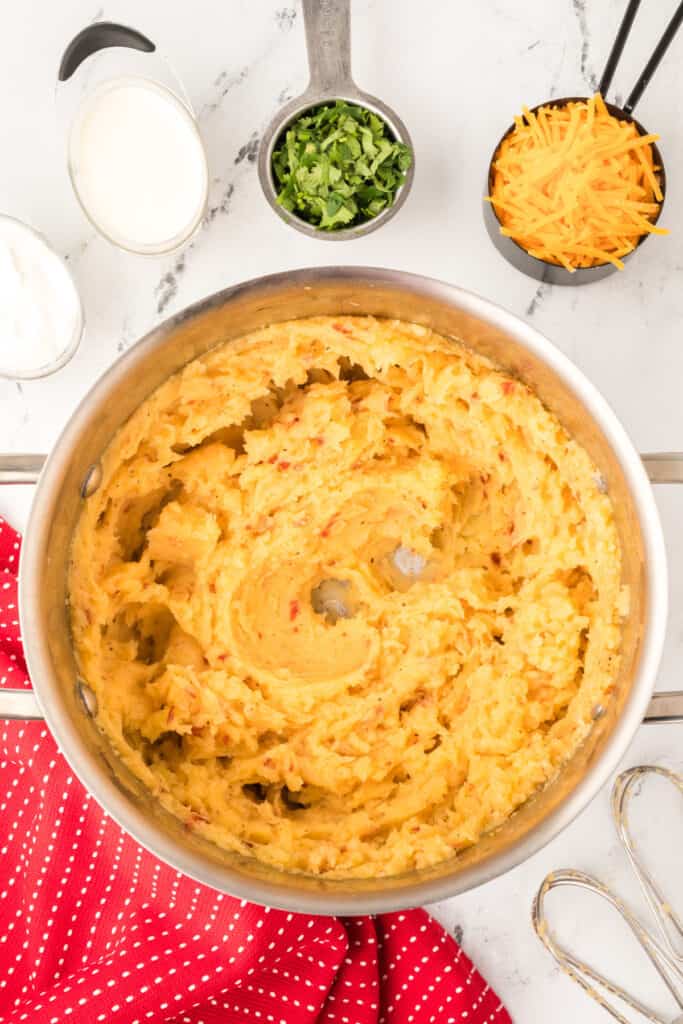 Mashed potatoes in a pot with milk, cilantro, and shredded Cheddar cheese nearby