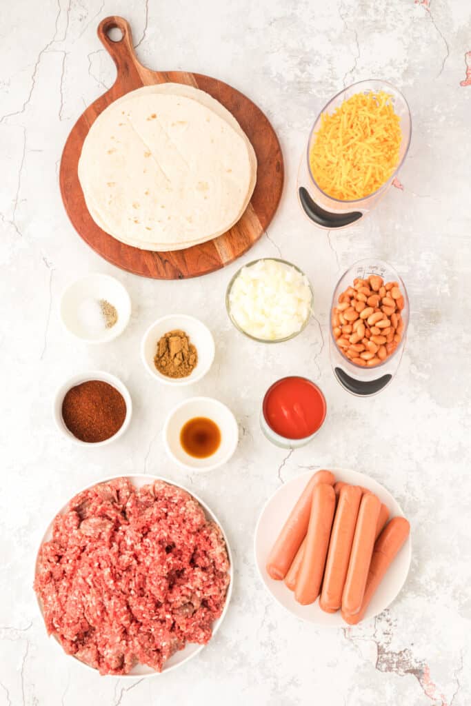 Ingredients for Chili Dog Casserole arranged on a light countertop, including flour tortillas, shredded Cheddar cheese, diced onion, pinto beans, tomato sauce, ground beef, hot dogs, and seasonings.