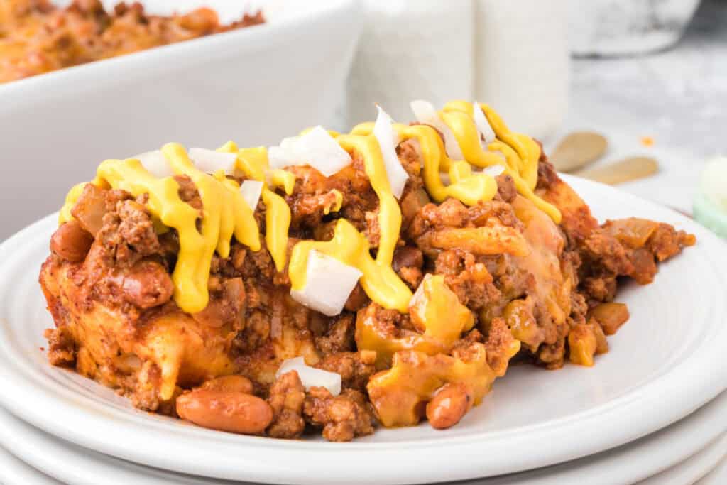 Slice of Chili Dog Casserole topped with yellow mustard and diced onions on a white plate with pinto beans, ground beef, melted cheese, and tortilla-wrapped hot dogs visible.