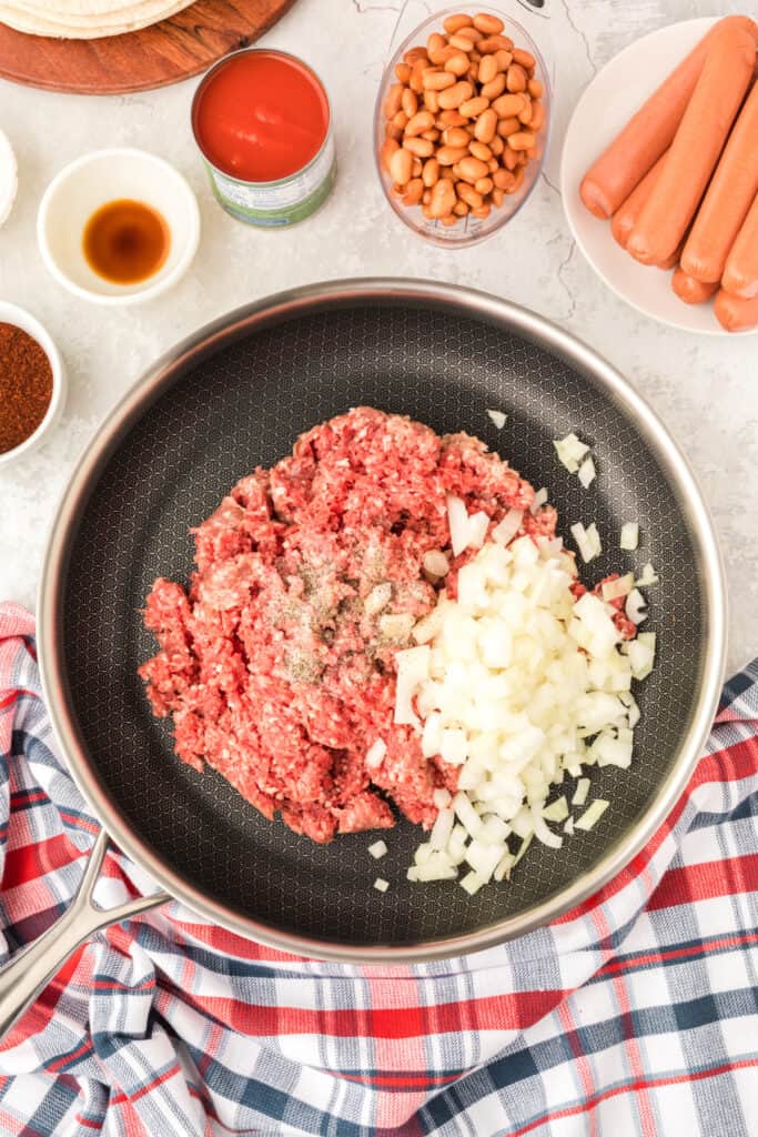 Raw ground beef, diced onion, salt, and pepper in a skillet with ingredients for Chili Dog Casserole arranged around the pan.