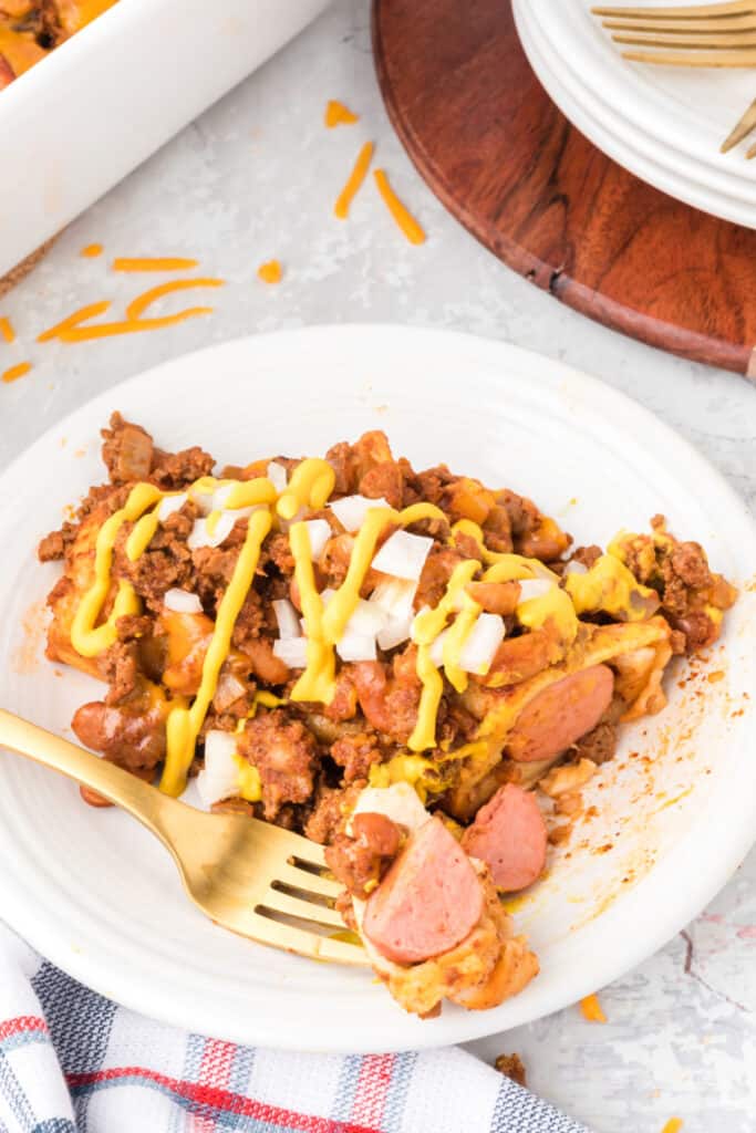 Close-up of a serving of Chili Dog Casserole on a white plate topped with yellow mustard and diced onions, showing sliced hot dogs, ground beef, pinto beans, and melted cheese.
