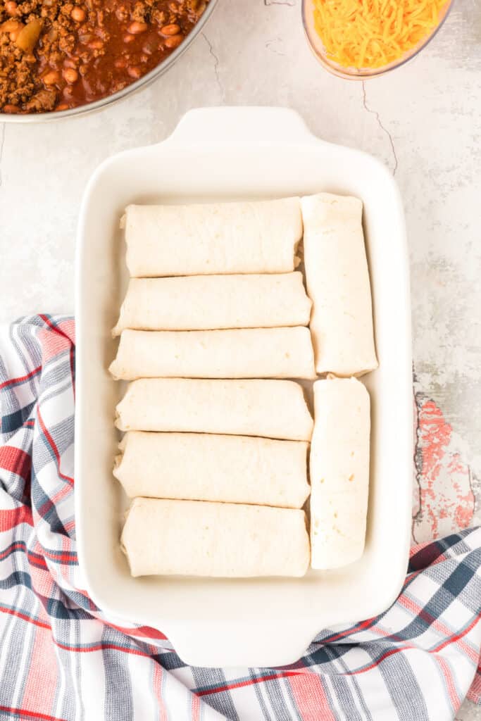 Hot dogs wrapped in flour tortillas arranged in a white baking dish before adding the chili mixture.