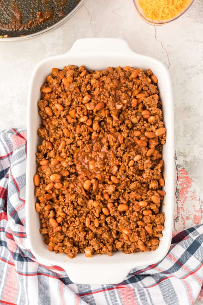 Chili mixture spread over tortilla-wrapped hot dogs in a white baking dish before adding cheese.