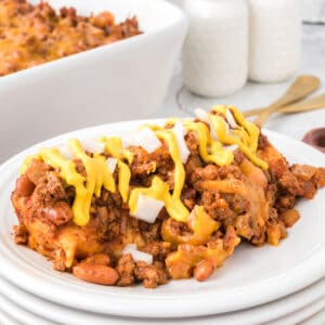 Square image of Chili Dog Casserole served on a white plate and topped with yellow mustard and diced onions, with visible ground beef, pinto beans, melted cheese, and tortilla-wrapped hot dogs.