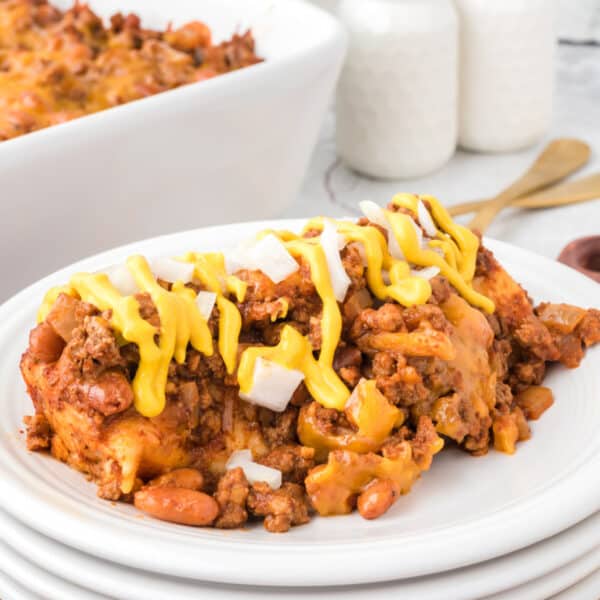 Square image of Chili Dog Casserole served on a white plate and topped with yellow mustard and diced onions, with visible ground beef, pinto beans, melted cheese, and tortilla-wrapped hot dogs.