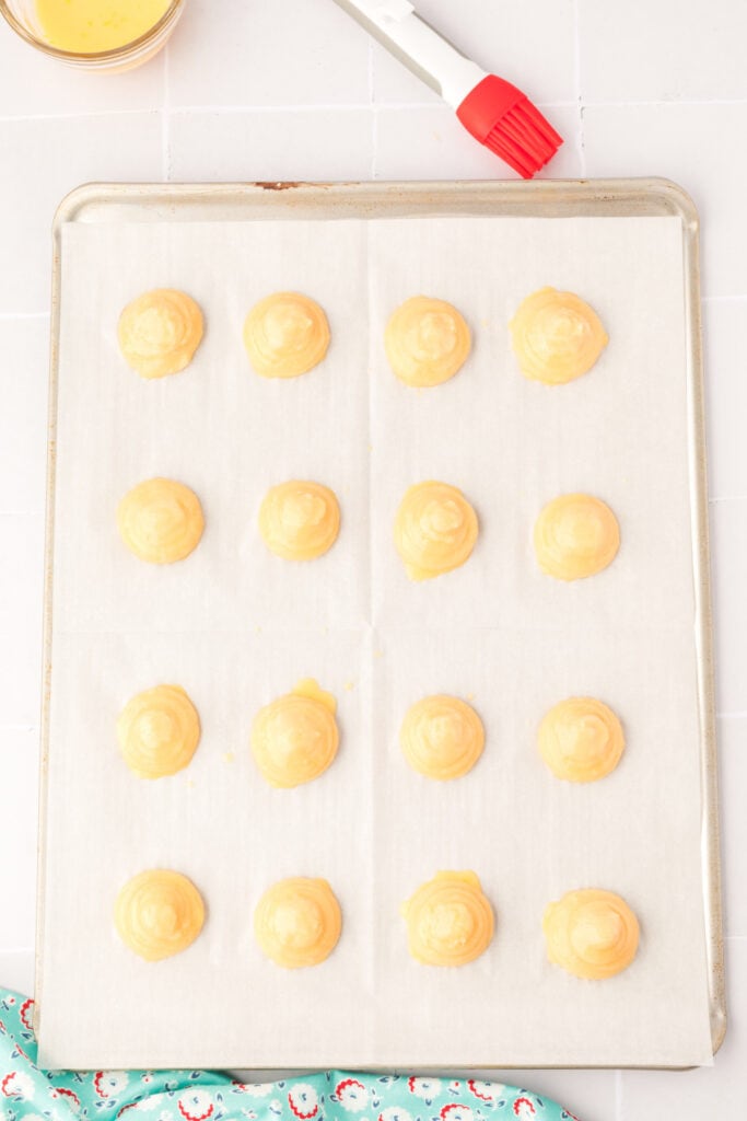 Pipe circles 1-½ inch round onto parchment paper. Moisten your finger with water and gently smooth out the pointed peaks, if necessary. Brush the pastry with egg wash.