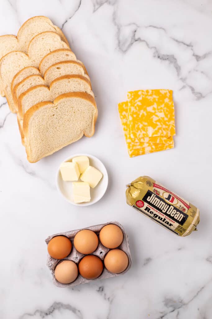 Ingredients for a Breakfast Grilled Cheese recipe including bread, eggs, sausage, butter, and cheese on a marble surface