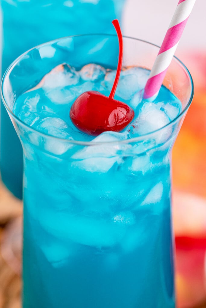 looking into a long tall glass with ocean water cocktail with a maraschino cherry and straw on top