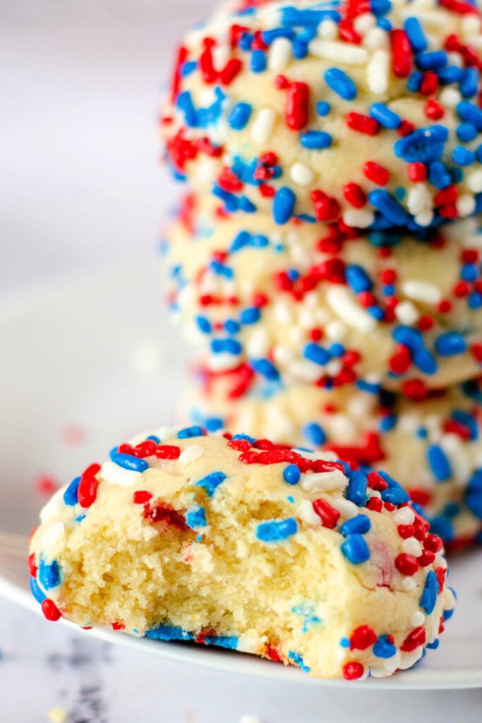 patriotic sprinkle cookies stacked in the background with the one in the front having a bite taken out of it