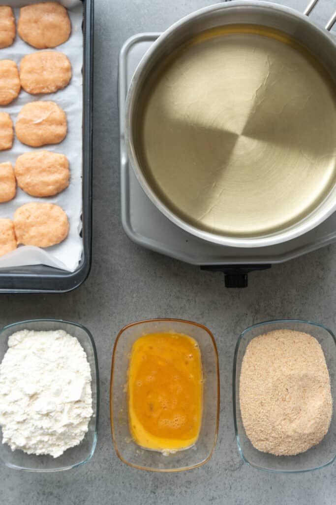 Breading station with flour, beaten eggs, seasoned breadcrumbs, and chilled homemade chicken nuggets ready for coating.
