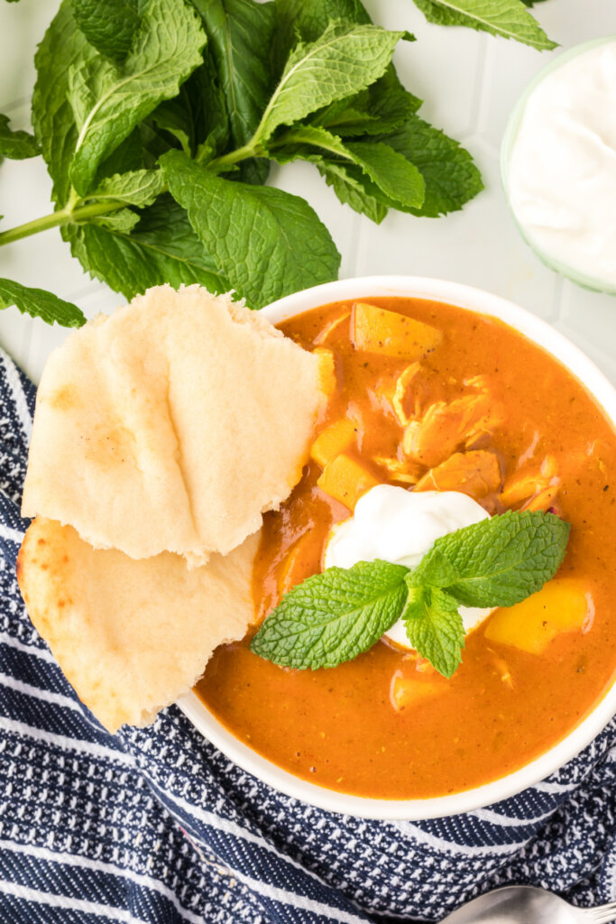 indian spiced chicken potato stew in white bowl garnished with yogurt mint and naan bread