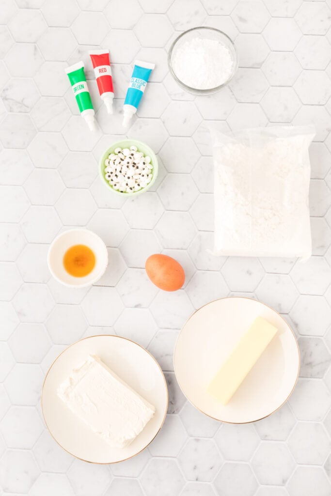 Ingredients for monster eye cookies including cake mix, cream cheese, butter, egg, vanilla, food coloring, and candy eyeballs laid out on a marble surface
