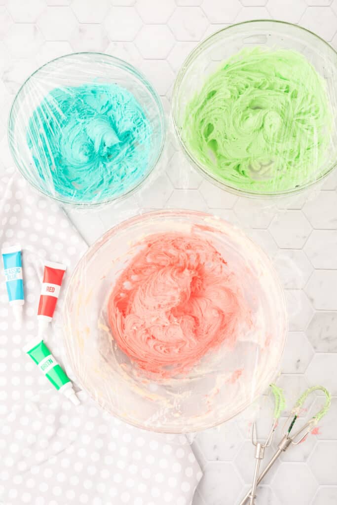 Three bowls of cookie dough tinted blue, green, and pink, covered with plastic wrap
