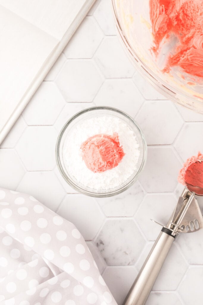 A ball of pink cookie dough dropped into a bowl of powdered sugar