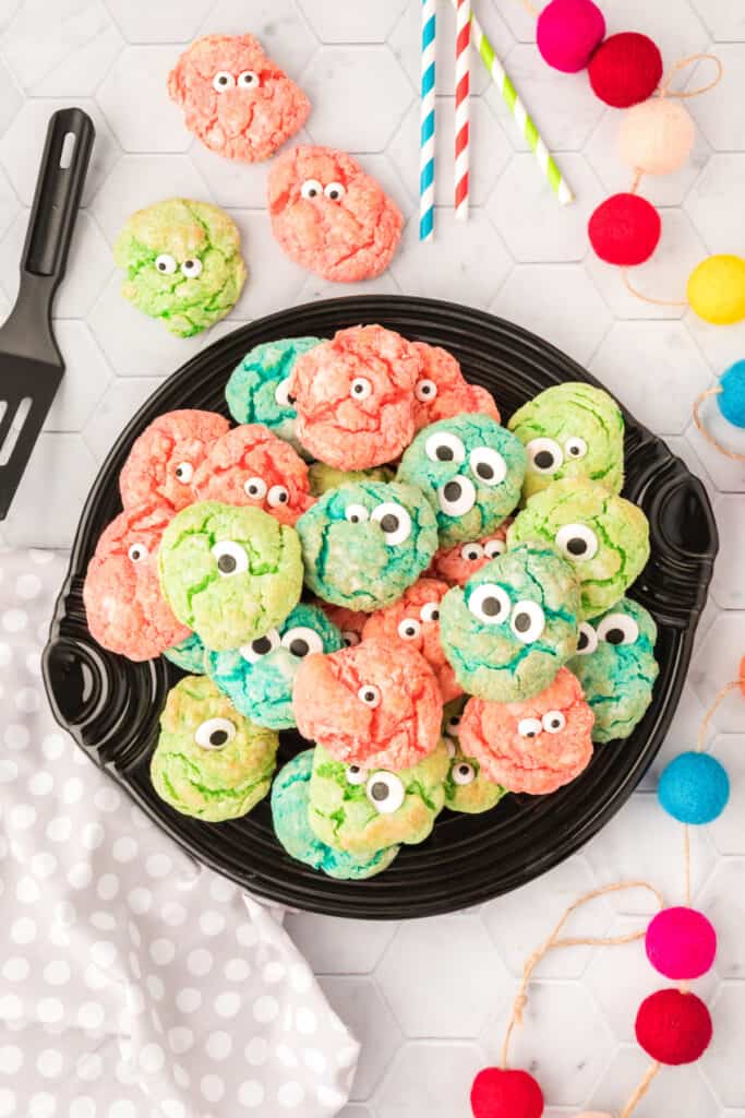 A black plate piled high with colorful pink, green, and blue monster eye cookies, surrounded by striped straws and pom pom garland