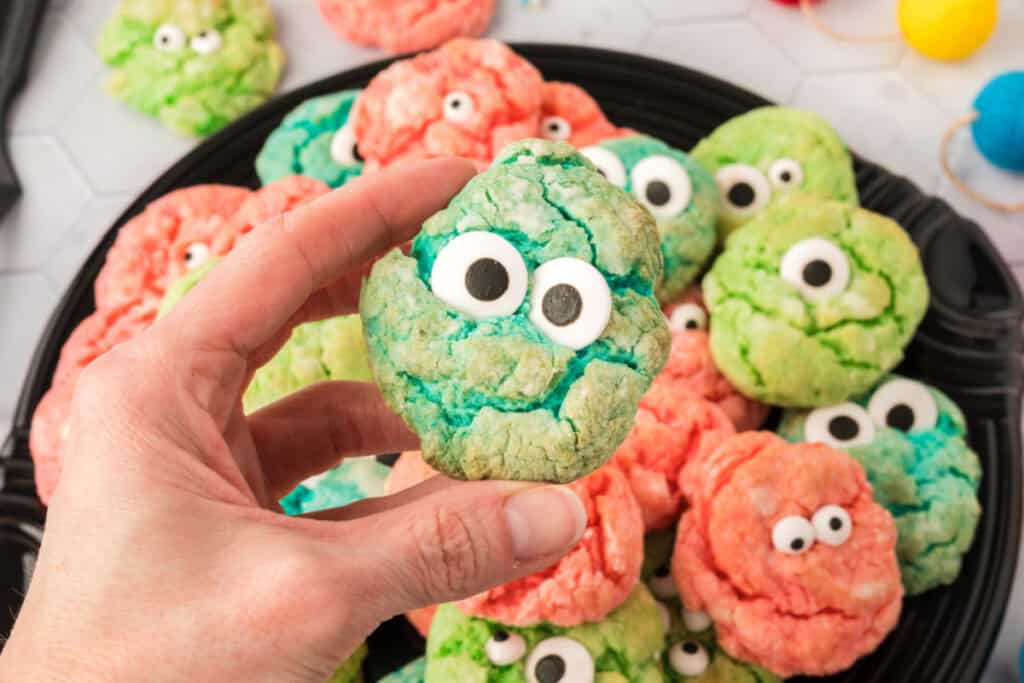 A hand holding up a teal monster eye cookie above a plate full of pink and green monster eye cookies