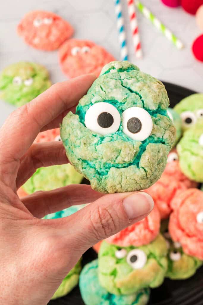 Close up of a hand holding a teal monster eye cookie with large candy eyes, with more cookies blurred in the background