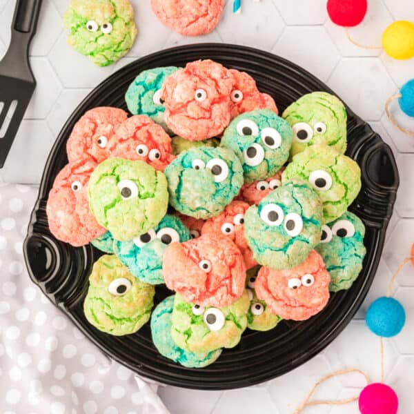 A square photo of a black plate piled with pink, green, and blue monster eye cookies decorated with candy eyes