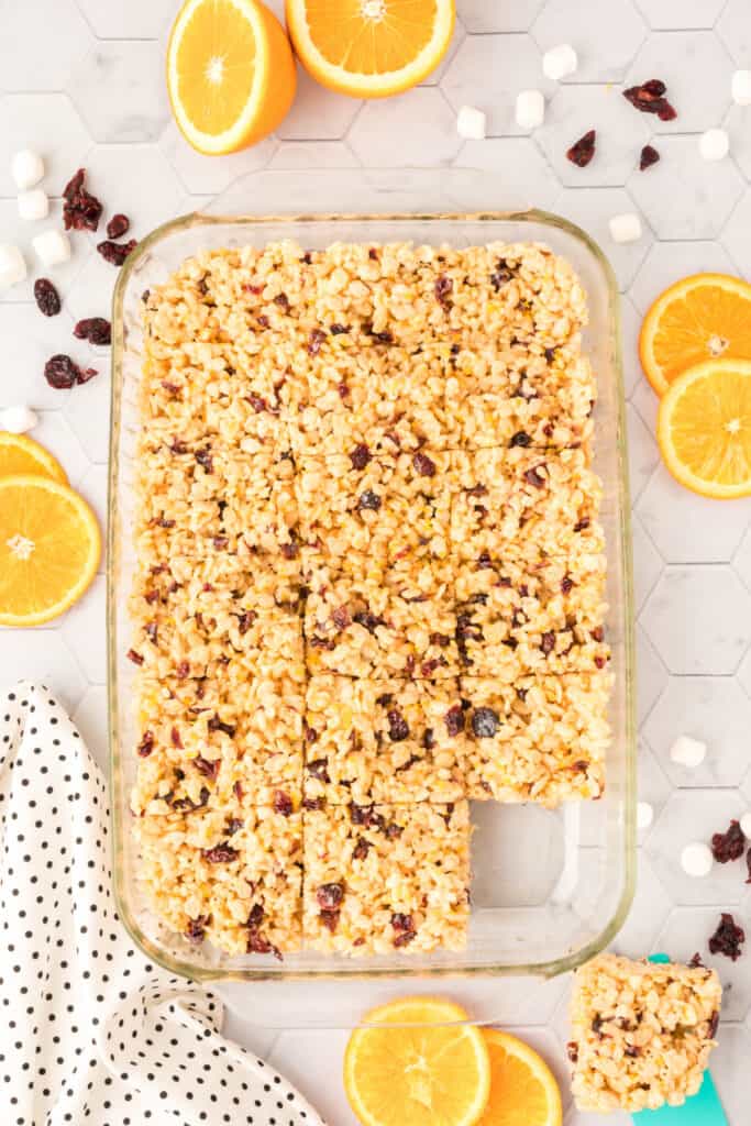 Pan of Cranberry Orange Rice Crispy Treats cut into squares and surrounded by oranges, marshmallows, and dried cranberries.