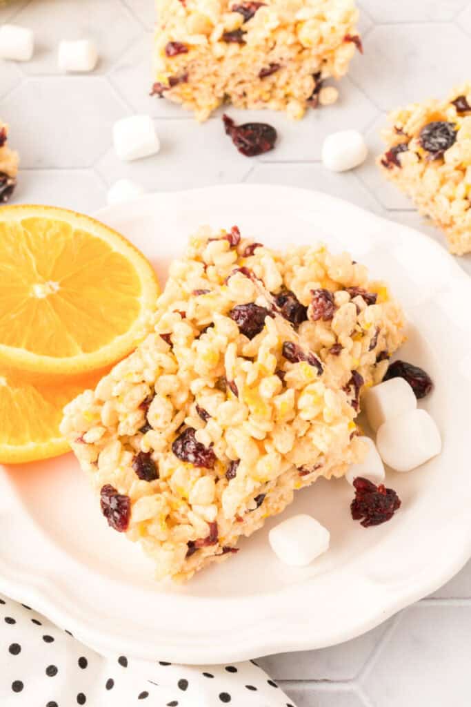 Two cranberry orange rice crispy treats served on a white plate with orange slices, marshmallows, and dried cranberries.