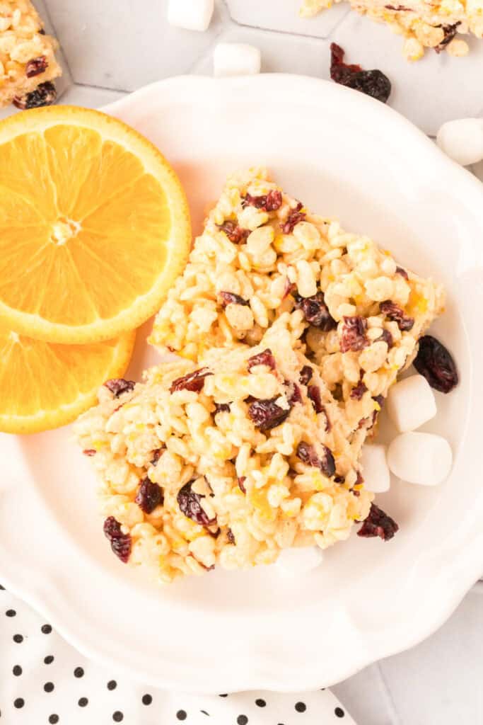Overhead view of cranberry orange crispy treat squares served with fresh orange slices and mini marshmallows.