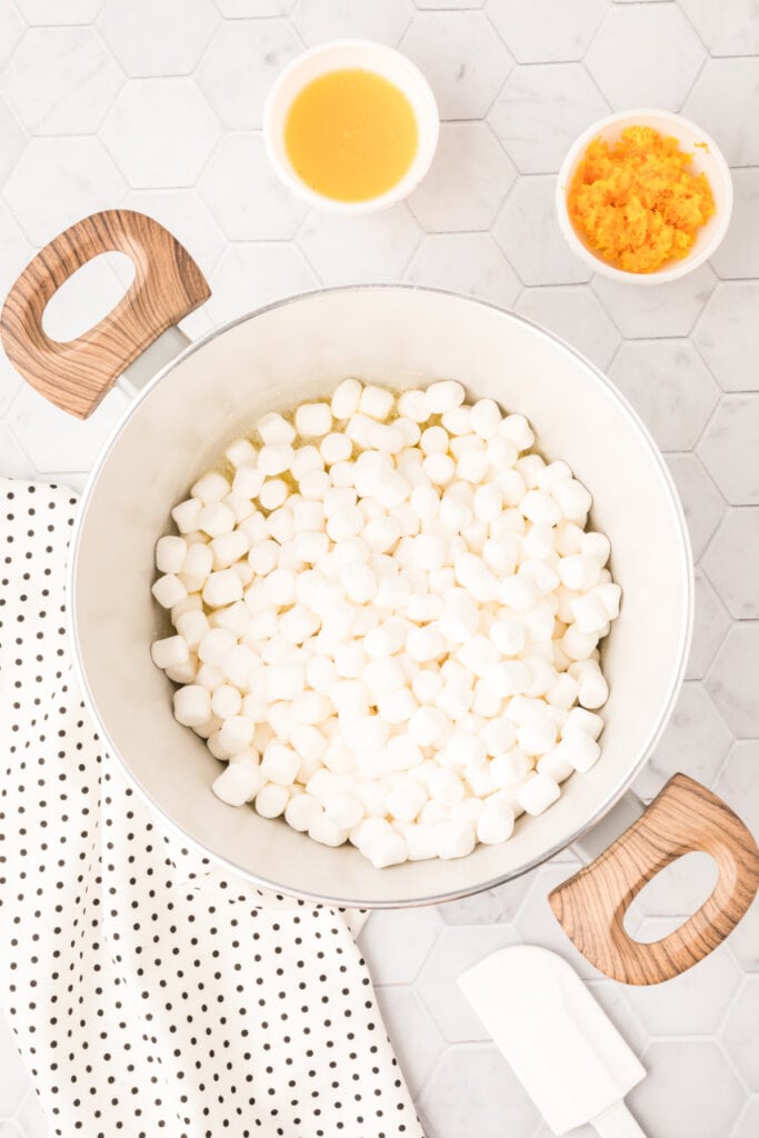Mini marshmallows added to melted butter in a large saucepan with orange juice and zest nearby.