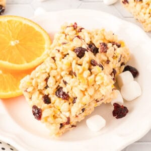 Cranberry Orange Rice Crispy Treats stacked on a plate with fresh orange slices, mini marshmallows, and dried cranberries.