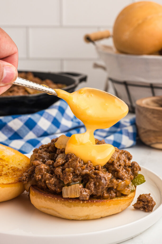 spooning melted cheese whiz onto the top of a philly cheesesteak sloppy joe