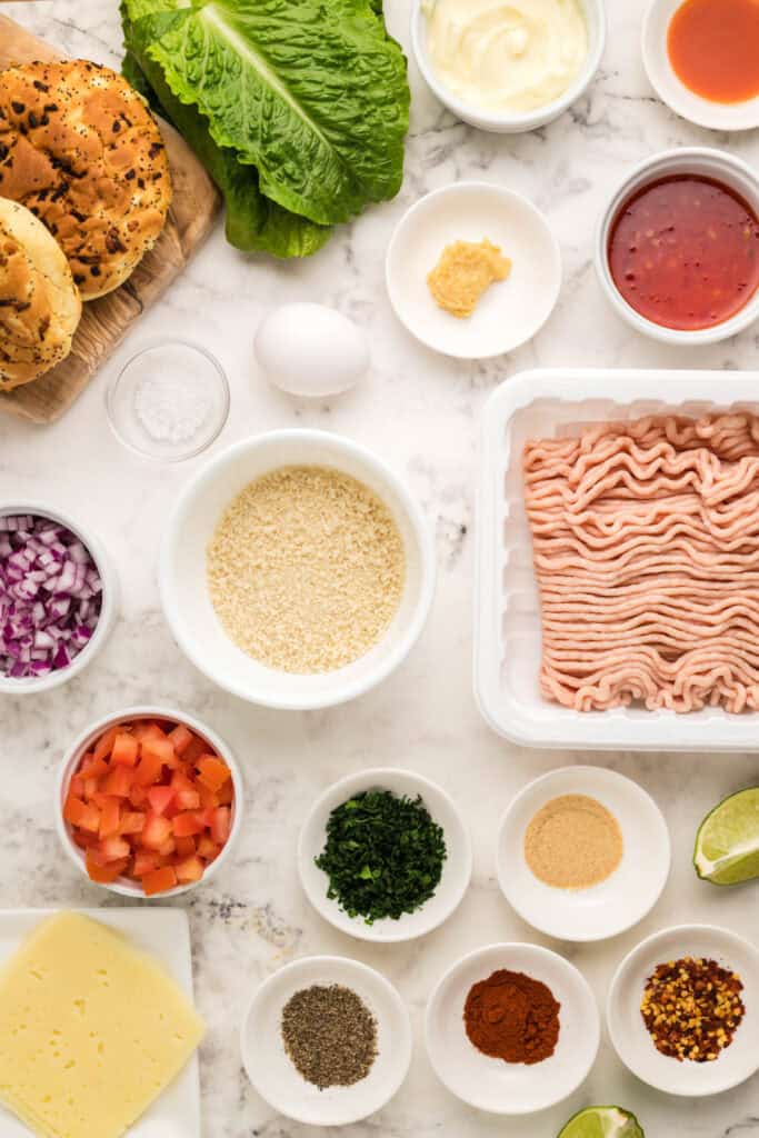 Ingredients for chicken burgers with bang bang sauce arranged on a marble countertop, including ground chicken, panko breadcrumbs, Swiss cheese, onion buns, romaine lettuce, diced tomato, red onion, cilantro, egg, lime, mayonnaise, sweet chili sauce, hot sauce, garlic, and seasonings.