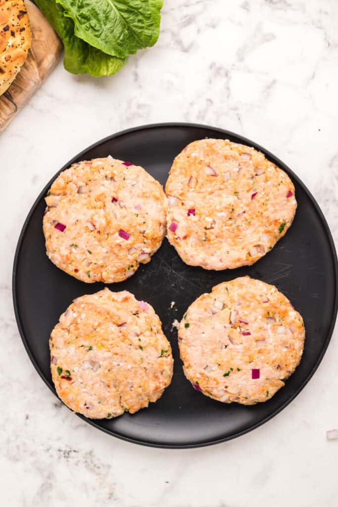 Four uncooked chicken burger patties shaped and ready for cooking on a black plate.