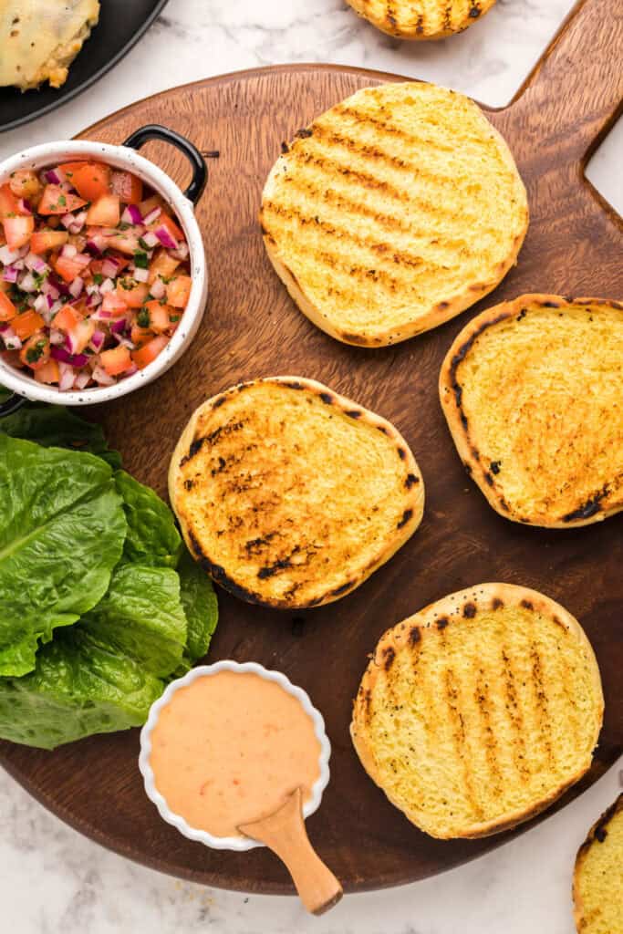 Toasted onion buns, fresh salsa, romaine lettuce, and bang bang sauce arranged on a wooden board.