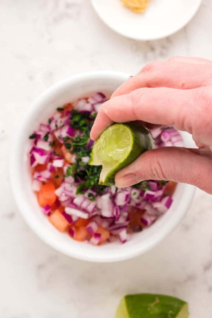 Hand squeezing fresh lime juice into a bowl of tomato, red onion, and cilantro salsa.
