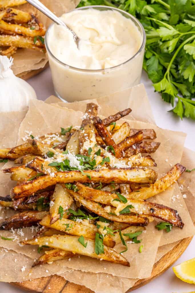 pile of air fryer garlic parmesan fries on parchment paper with a bowl of garlic aioli in background