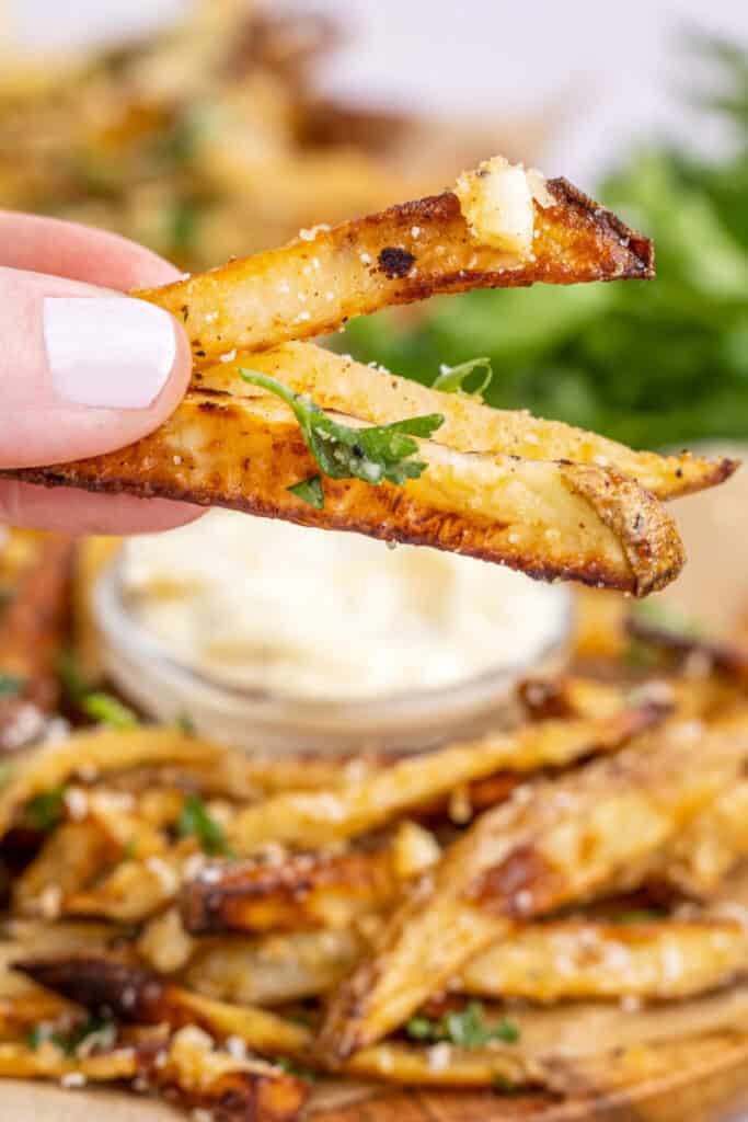 hand holding a few fries over a plate of fries with a jar of garlic aioli