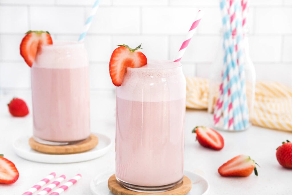 two glasses filled with strawberry milk with a glass filled with striped straws in the back right