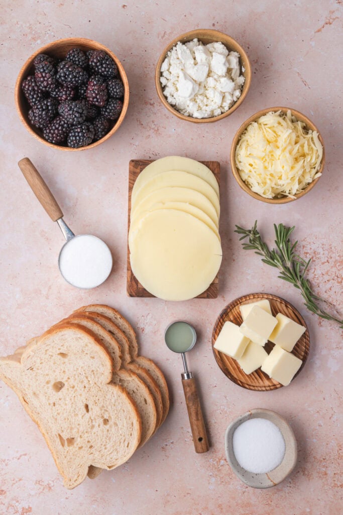 Flat lay of all ingredients for blackberry grilled cheese including blackberries, cheeses, butter, bread, rosemary, and sugar