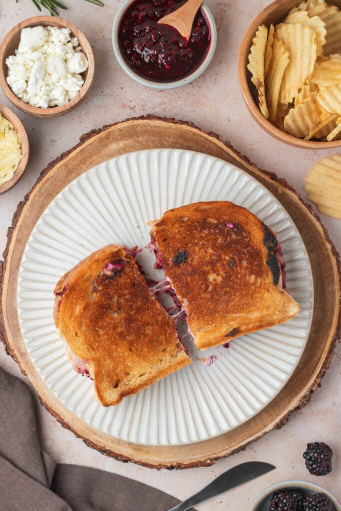 Halved blackberry grilled cheese sandwich on a plate with chips, jam, and cheese bowls